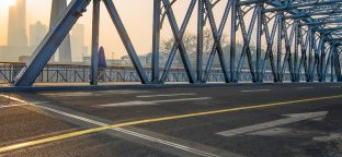 A large empty bridge with three traffic lanes with a modern city skyline in the background at sunset.