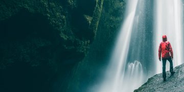 A low-angel view of a person standing on a boulder looking up at a waterfall.