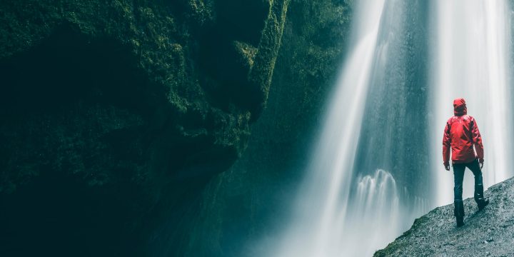 A low-angel view of a person standing on a boulder looking up at a waterfall.