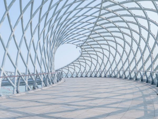 A close-up view of a futuristic pedestrian bridge with a metal crisscross tunnel-archway against the backdrop of a blue sky.