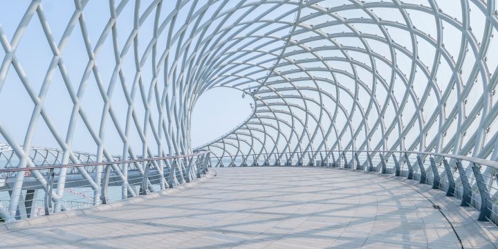 A close-up view of a futuristic pedestrian bridge with a metal crisscross tunnel-archway against the backdrop of a blue sky.
