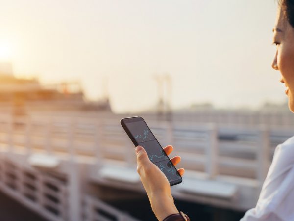 A businesswoman checking financial data on a smartphone in a commercial dock in a city at sunset.