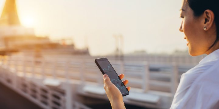 A businesswoman checking financial data on a smartphone in a commercial dock in a city at sunset.